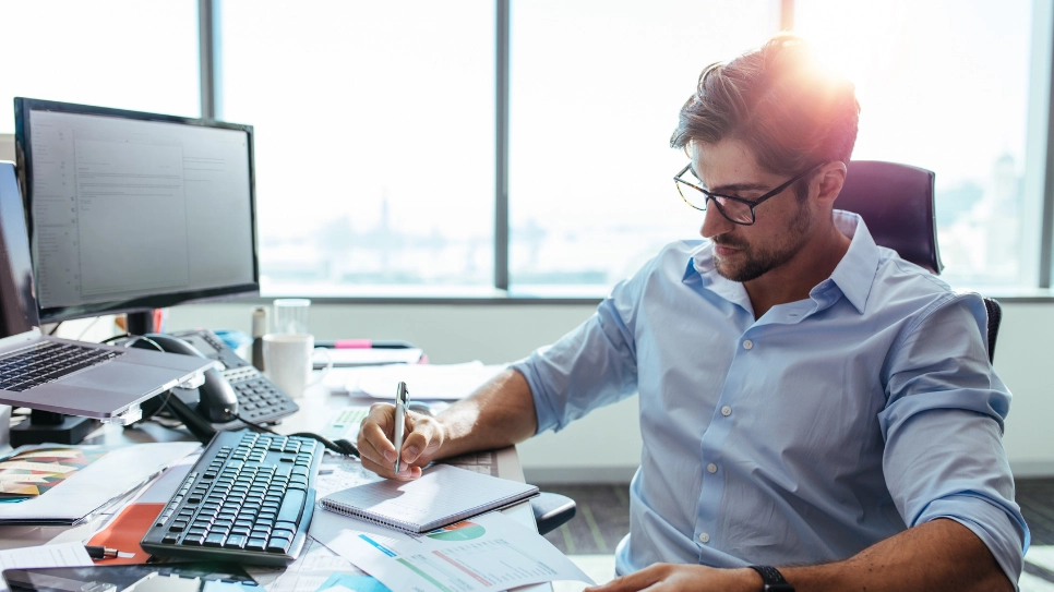 Businessman is writing in his notes sitting behind his desk with a computer.