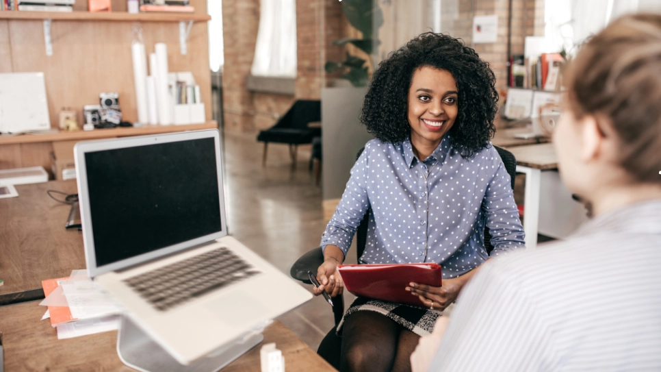 A smiling HR professional conducts an interview, holding a notepad in a modern office setting.