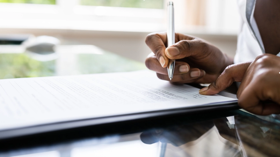 Close-up of a hand holding a pen, signing a legal document on a reflective glass table, symbolizing legal agreements or contracts.