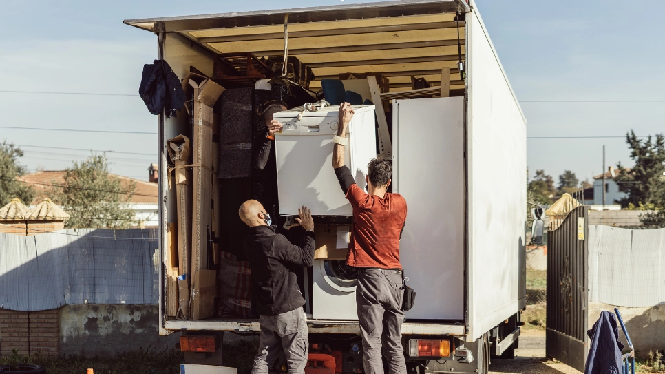 Two movers carefully loading a washing machine and household items into a moving truck for transportation.