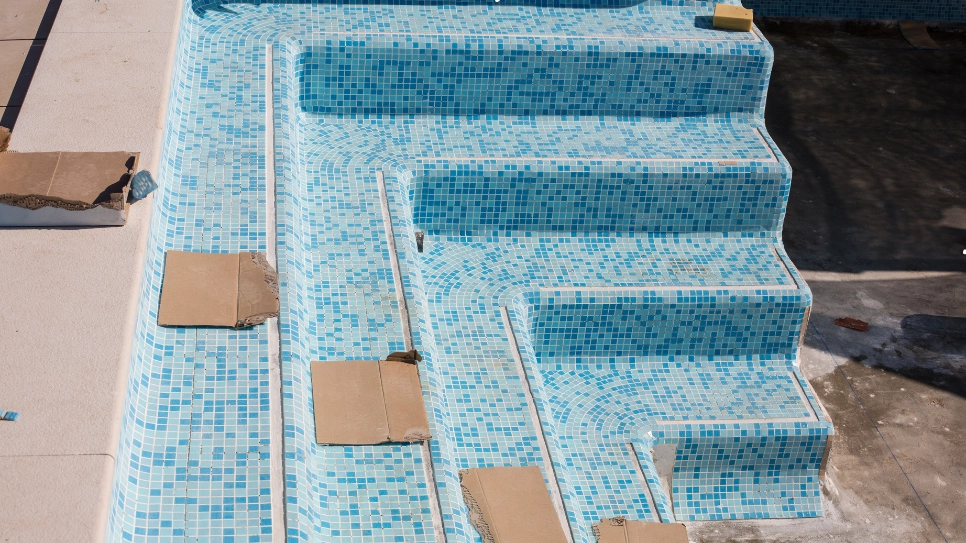 Installation of blue mosaic tiles on a swimming pool staircase, showcasing detailed craftsmanship and design.