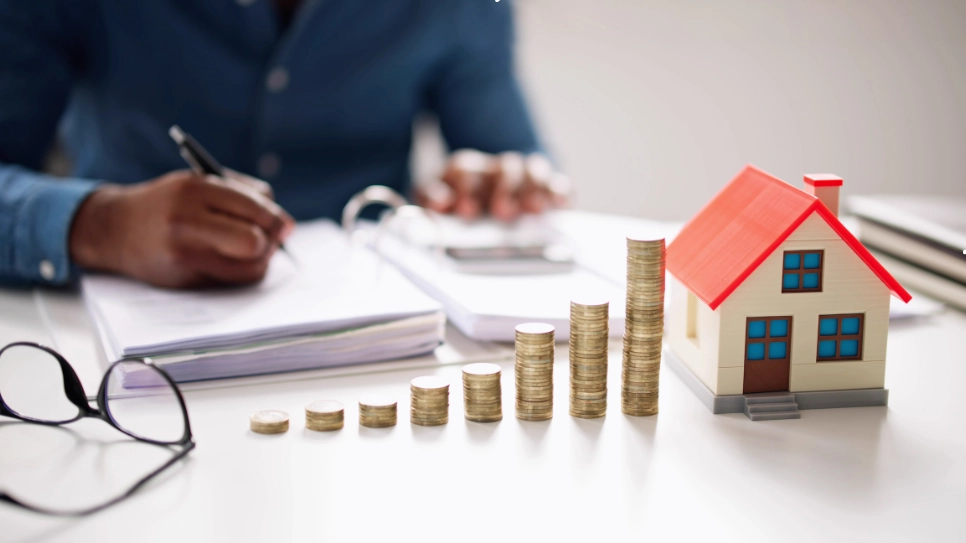 Stacked coins increasing in size next to a model house, with a person calculating finances in the background.