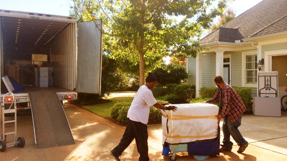 Two movers carefully unloading furniture wrapped for protection outside a residential home with a moving truck.