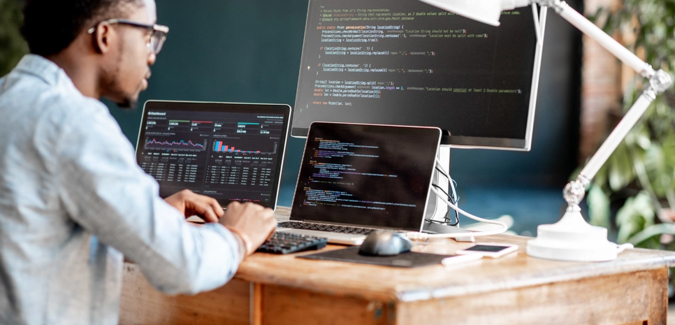 Young african programmer writing computer code sitting in a workplace with three monitors.