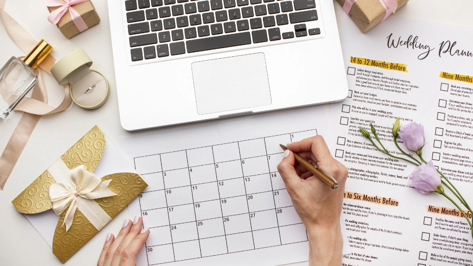 A flat lay of wedding planning essentials, featuring a checklist timeline, calendar, and a laptop. The scene is decorated with pastel flowers, ribbon, small gift boxes, and a golden envelope, embodying a well-organized and aesthetically pleasing wedding planning setup. The timeline lists important tasks for different months leading up to the wedding, helping couples stay on track for their big day.