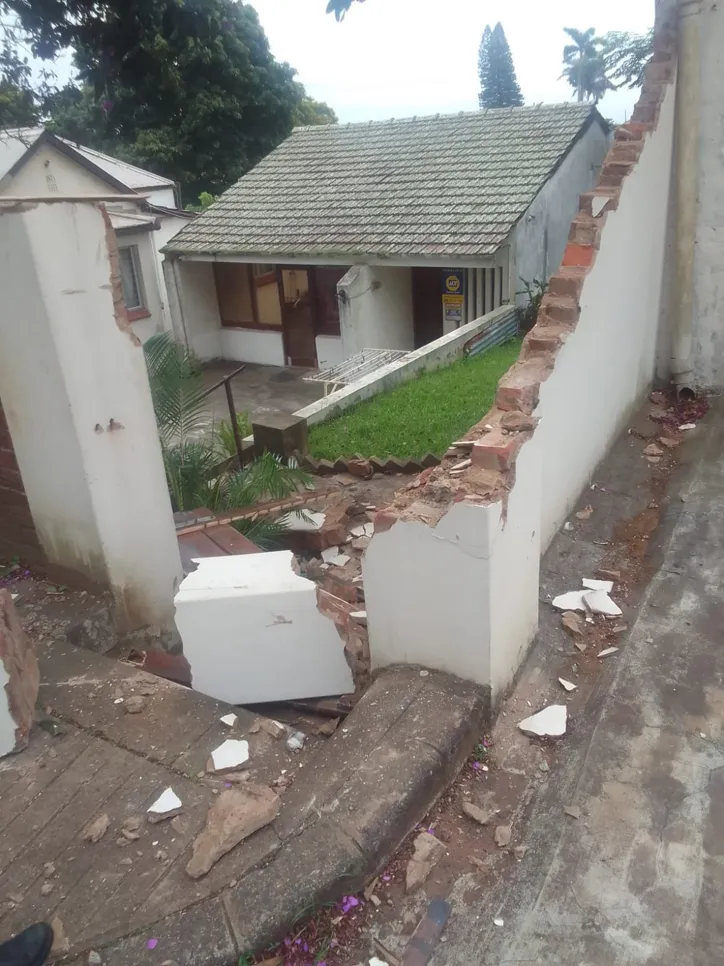 Damaged brick wall in residential area Collapsed brick wall in front of a house, showing structural damage and debris on the ground.